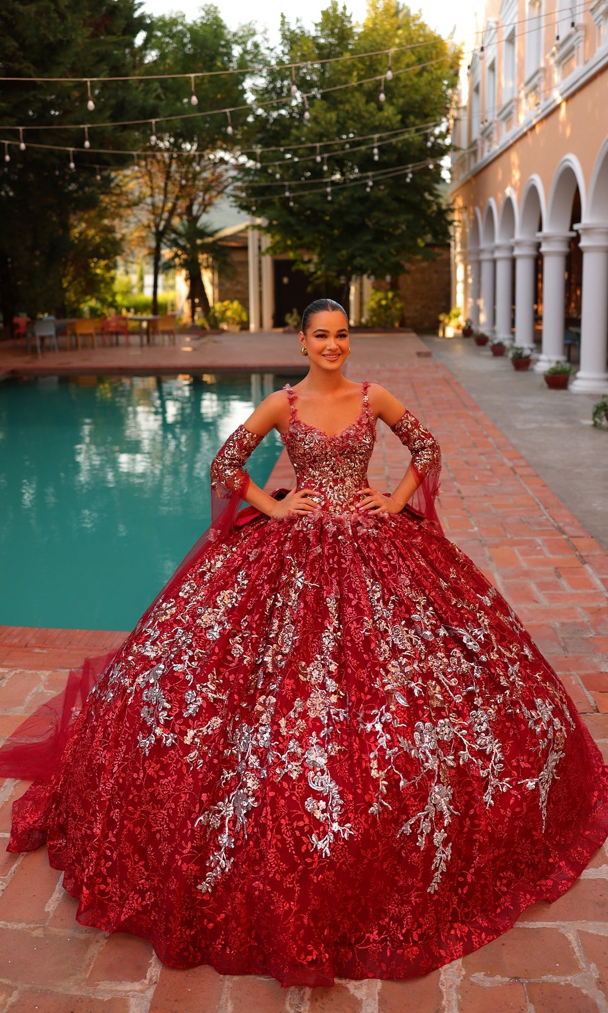A woman stands on a brick patio by a pool, wearing the Quinceanera Dress 54322 By Amarra?a stunning floral embroidered ball gown with matching long sleeves. Trees and an arched building provide an elegant backdrop.